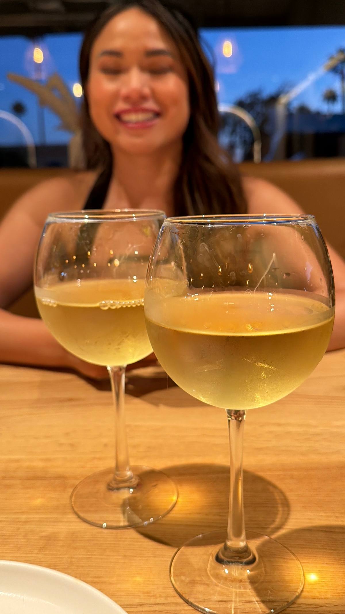 Two glasses of chilled chardonnay on a wood table, Virginia smiling behind them in the warm-lit dining room.