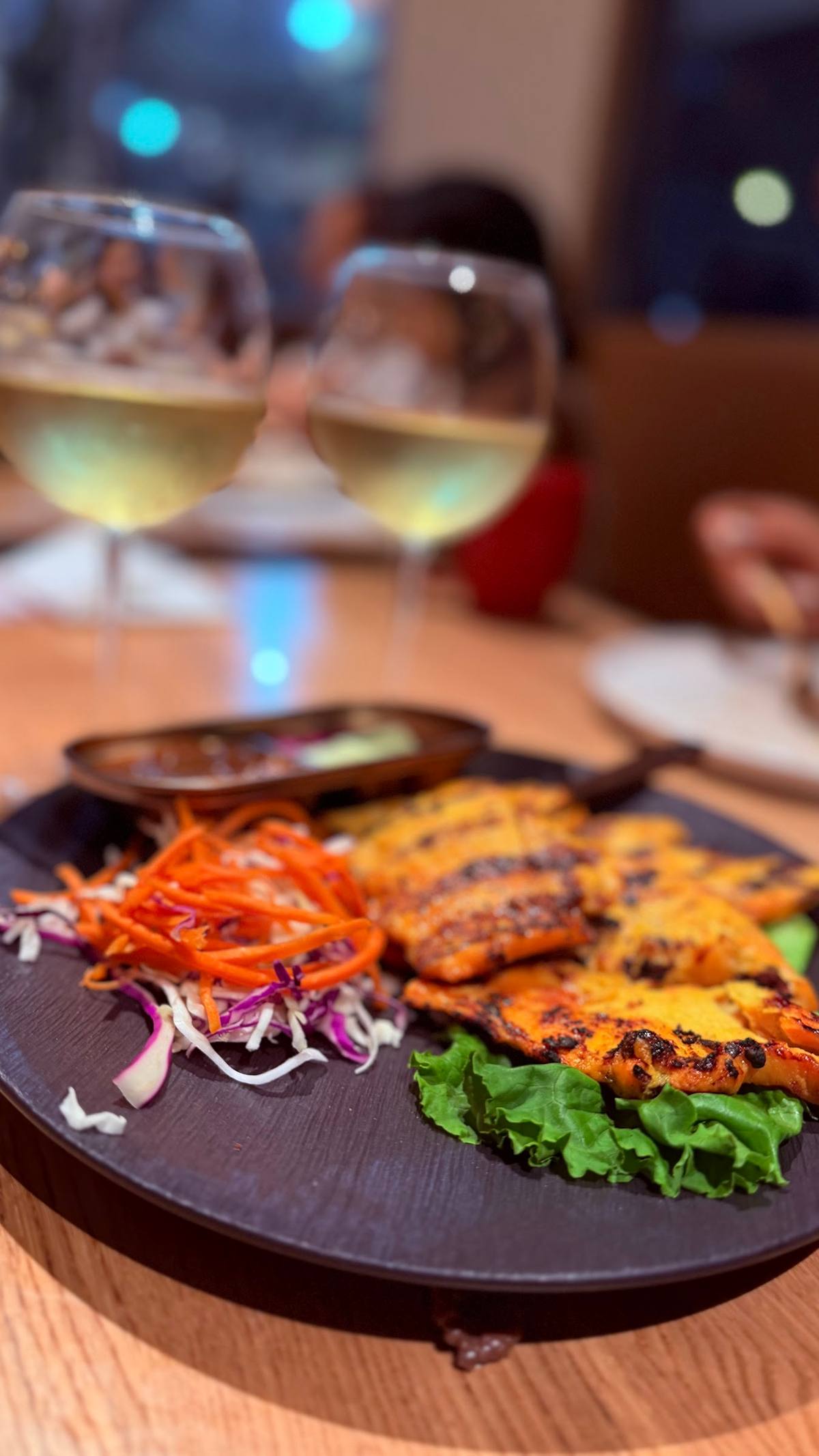 Grilled turmeric chicken on a dark plate with slaw and dipping sauces, two glasses of chardonnay glowing behind it.