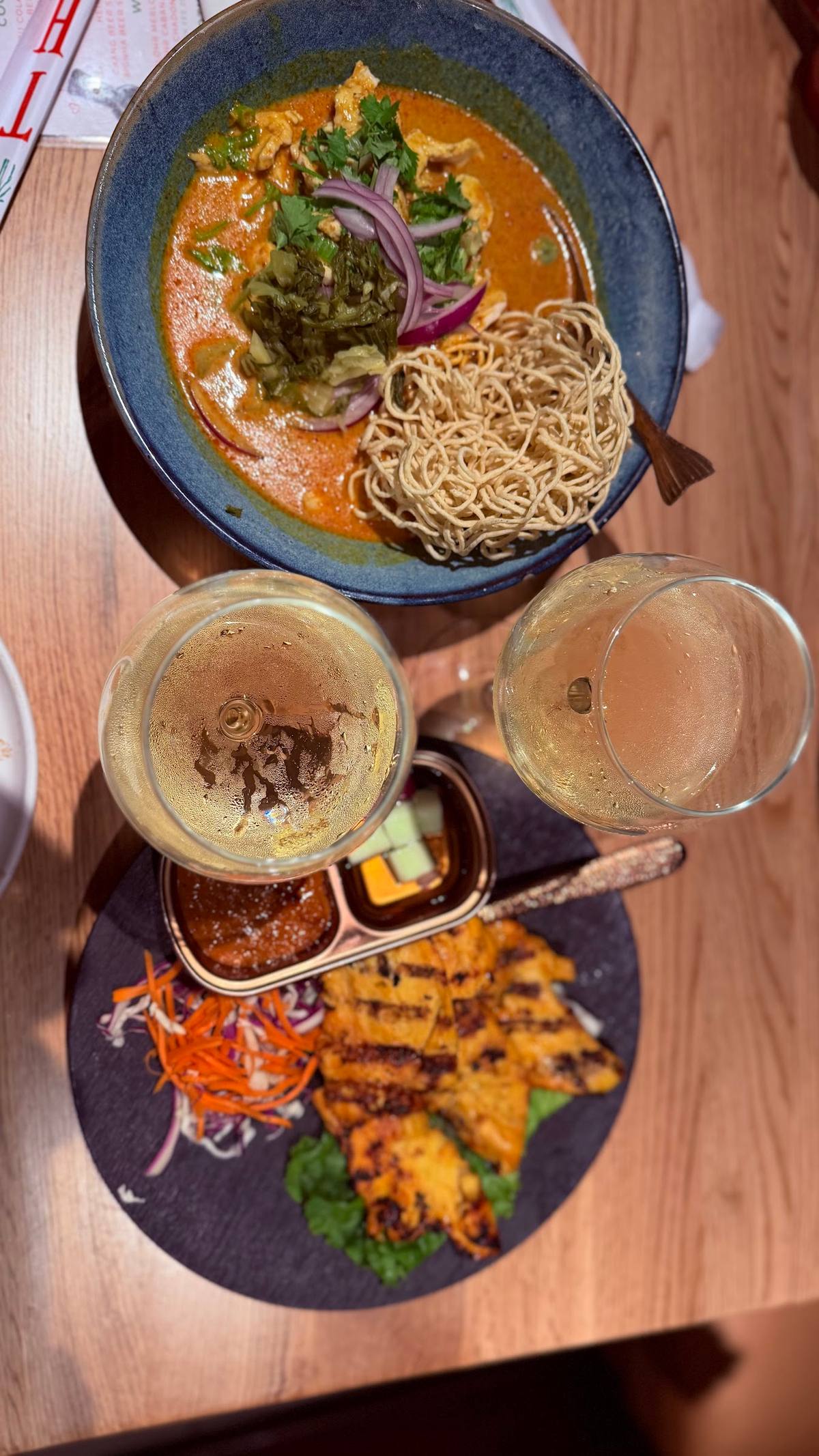 Overhead of the table: a blue bowl of khao soi, a plate of grilled chicken with slaw and dipping sauces, and two glasses of chardonnay.