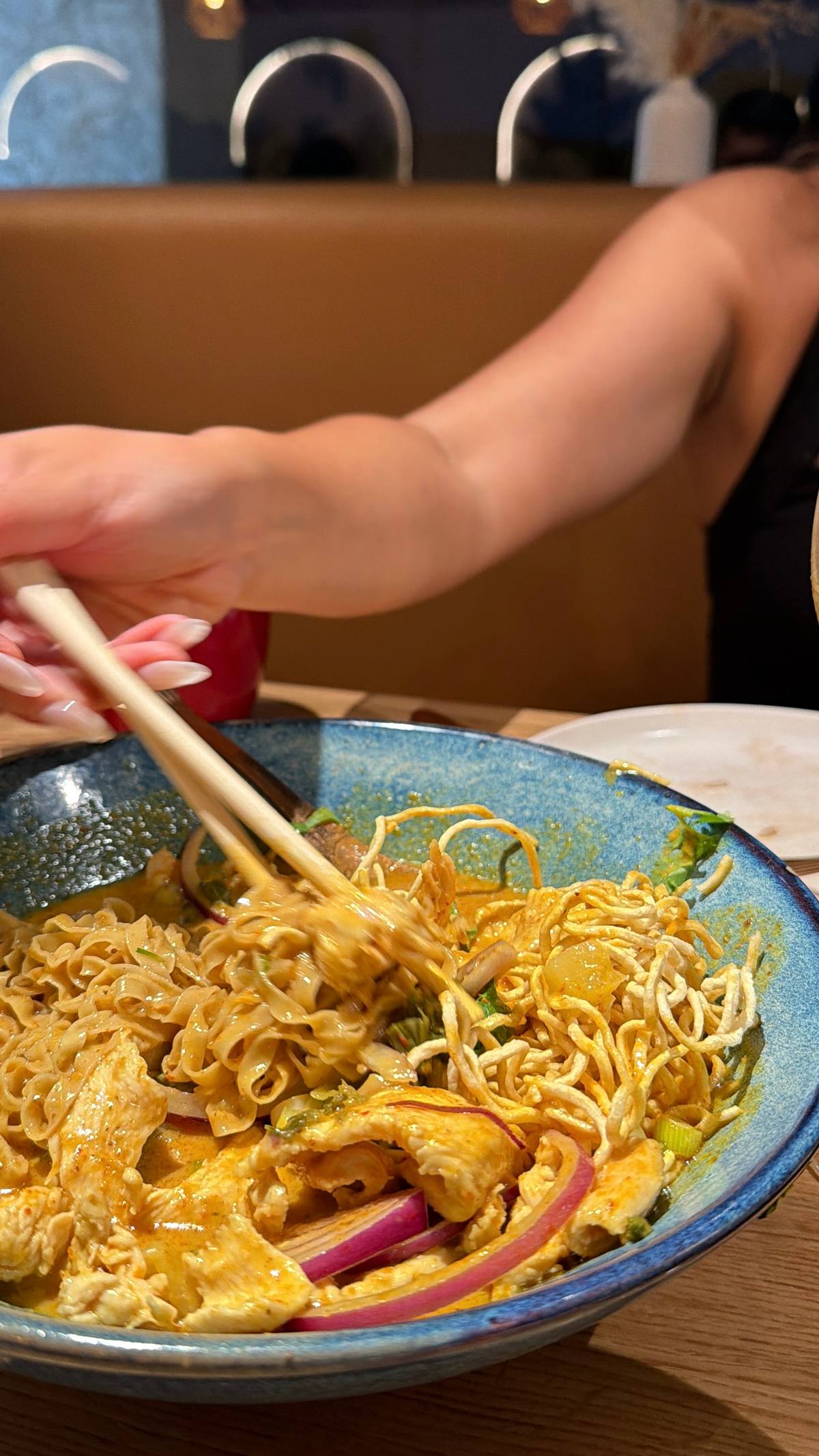 Close-up of khao soi in a speckled blue bowl: golden coconut curry, soft egg noodles, a nest of crispy noodles, sliced chicken, pickled red onion and herbs, chopsticks pulling a bite.