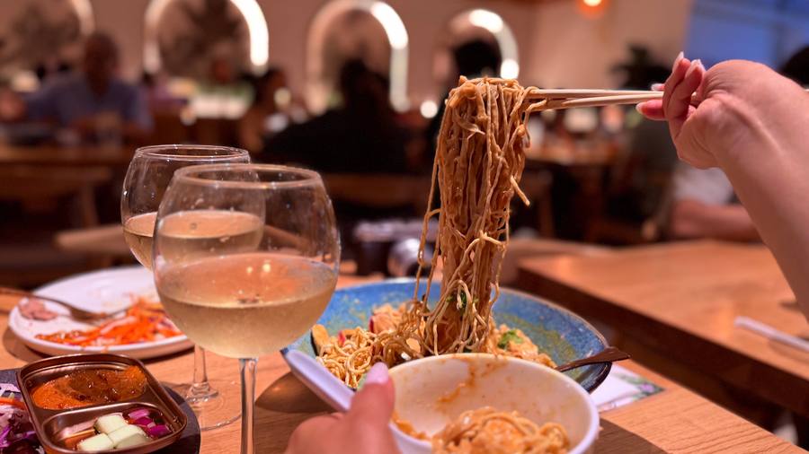 A high pull of golden egg noodles lifted on chopsticks from a bowl of khao soi, two glasses of chardonnay in the foreground, the warm modern dining room behind.
