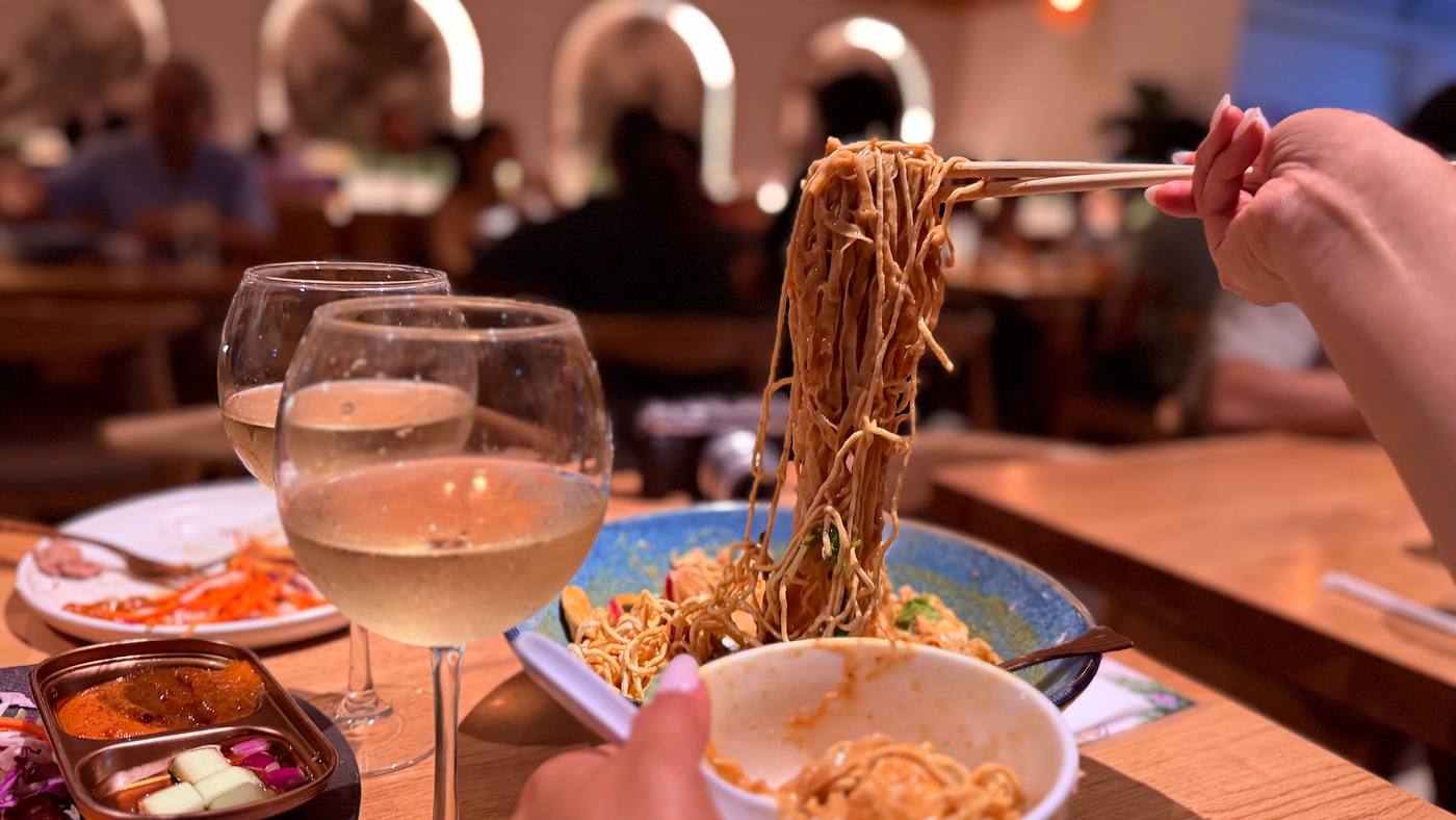 A high pull of golden egg noodles lifted on chopsticks from a bowl of khao soi, two glasses of chardonnay in the foreground, the warm modern dining room behind.